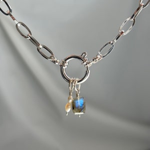 May include: A silver chain necklace with a large oval link design. The necklace has a toggle clasp and a charm with a blue labradorite stone and a pearl.