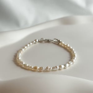 Peut inclure: Un bracelet délicat composé de petites perles blanches de forme irrégulière. Le bracelet est doté d'un fermoir argenté. Les perles sont enfilées ensemble, créant un bijou fin. Le bracelet est présenté sur une surface blanche.