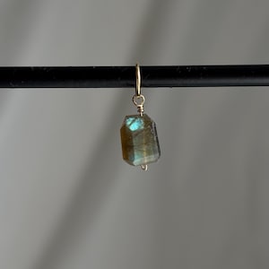 Puede incluir: Un solo pendiente dorado con una gema de labradorita facetada y rectangular. La piedra muestra tonos iridiscentes azules y verdes. El pendiente está suspendido de un fino alambre dorado.