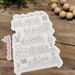 May include: A white sticker with a bookshelf design, featuring the words "Books of the Bible" at the top. The sticker includes the names of the books of the Bible, arranged as if they were on shelves, with potted plants. A small sticker with the text "@MakerInSurprise" is also present.