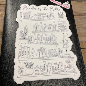 May include: A black Bible with a white sticker featuring a bookshelf design. The sticker lists the books of the Bible in a decorative font, with small potted plants. The sticker has the text "Books of the Bible".