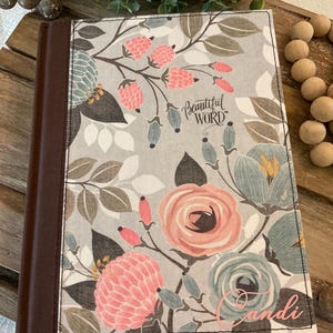 May include: A floral-patterned book cover with a brown spine. The cover features pink and blue flowers, berries, and the text "Beautiful Word." The name "Candi" is written in pink script on the bottom right corner. The background is a light gray.