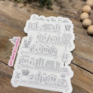 May include: A white sticker featuring a bookshelf design with the words "Books of the Bible" at the top. The shelves are filled with book titles and small potted plants. The sticker has a black outline and is on a wooden surface.