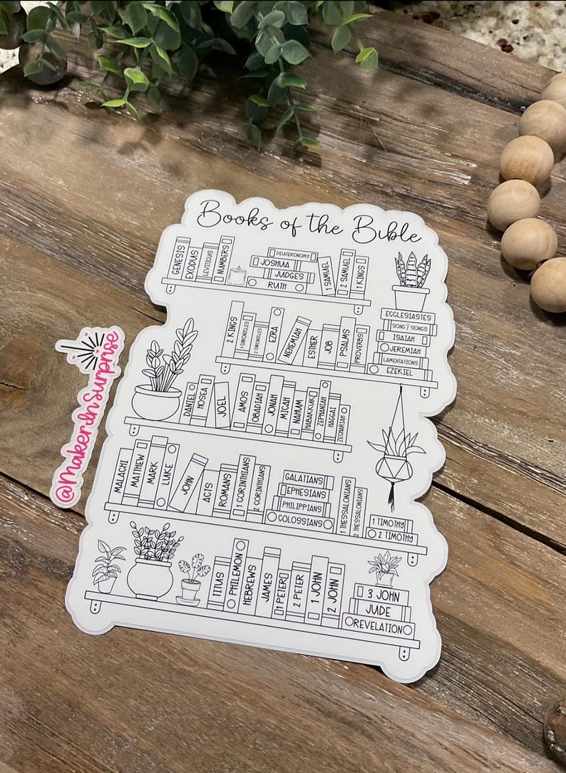 May include: A white sticker featuring a bookshelf design with the words "Books of the Bible" at the top. The shelves are filled with book titles and small potted plants. The sticker has a black outline and is on a wooden surface.