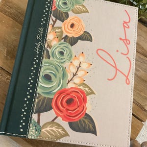 May include: A personalized Bible with a floral design. The cover features a light gray background with a colorful floral arrangement of roses in shades of red, peach, and green. The name "Lisa" is written in coral script.