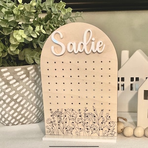May include: Wooden earring holder with the name Sadie on top. The holder has a floral design at the bottom and a grid of holes for earrings.