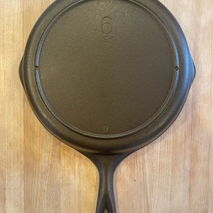 May include: A cast iron skillet with a black finish and a single loop handle. The skillet has a 6" diameter and is marked with the letter "D".