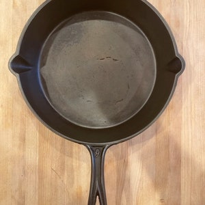 Puede incluir: Una sartén de hierro fundido con un mango negro. La sartén tiene un diámetro de 23 cm y una superficie lisa y negra.