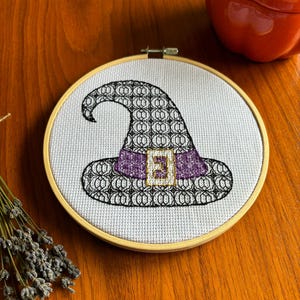 May include: A cross-stitch embroidery of a witch's hat in a wooden hoop. The hat is black with a repeating pattern, a purple band, and a gold buckle. The background is white fabric.
