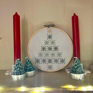 May include: A Christmas-themed display featuring a cross-stitch hoop art with a snowflake tree design, flanked by two tall red candles and three small green tree-shaped candles. The candles are on a white surface.