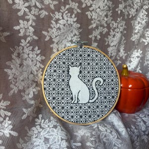 May include: A circular wooden embroidery hoop containing a white fabric piece with a black cross-stitched silhouette of a cat centered against a dense, repeating black floral pattern. An orange ceramic pumpkin sits to the right of the hoop.