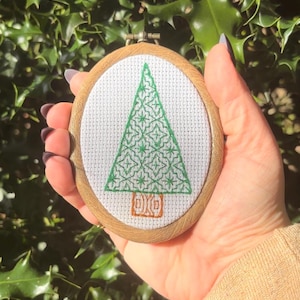 Puede incluir: Un pequeño bastidor de bordado ovalado con un diseño de árbol de Navidad verde sobre tela blanca. El árbol presenta patrones intrincados y un tronco marrón. El bastidor de madera añade un toque rústico a la decoración navideña hecha a mano.