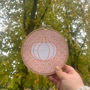 May include: A round cross-stitch embroidery hoop featuring an orange pumpkin design. The pumpkin is outlined in orange thread against a white background. The background has an orange pattern. The hoop is held in front of a blurred green and yellow tree.