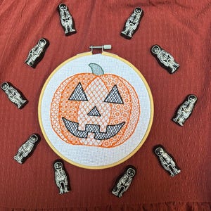 Puede incluir: Un bastidor de bordado con temática de Halloween con un diseño de calabaza naranja y una cara negra, rodeado de pequeños alfileres de esqueleto en blanco y negro. El bastidor se encuentra sobre un fondo de tela color óxido, creando una exhibición festiva de otoño.