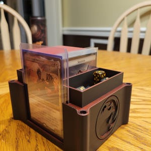 Può includere: Un organizzatore per giochi di carte stampato in 3D di colore nero e rosso con un logo di drago. L'organizzatore ha due scomparti per le carte e uno scomparto separato per i dadi. Gli scomparti per le carte hanno buste di plastica trasparente per contenere le carte.