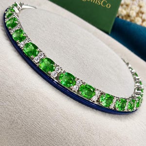 Op de afbeelding: Een zilverkleurige tennisarmband met een herhalend patroon van ovaal geslepen, levendig groene edelstenen afgewisseld met paren kleine, heldere, rond geslepen stenen. De armband ligt op een gebogen oppervlak naast een groen doosje met het opschrift DionagemsCo.