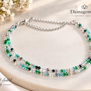 May include: A double-strand anklet featuring faceted crystal beads in shades of emerald green, turquoise, black, and clear white. The jewelry piece includes a silver-toned metal chain with a lobster claw clasp and an adjustable extender chain.