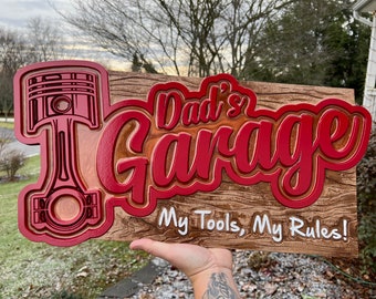 May include: Red wooden sign with the text "Dad's Garage" in a red 3D font. The sign also has a red 3D image of a piston and the text "My Tools, My Rules!" in white.