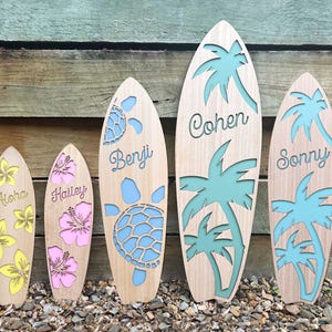 May include: Five wooden surfboard-shaped ornaments with personalised names. The ornaments are painted in various colours, including yellow, pink, and blue. Each ornament features a cutout design of a palm tree or a turtle.