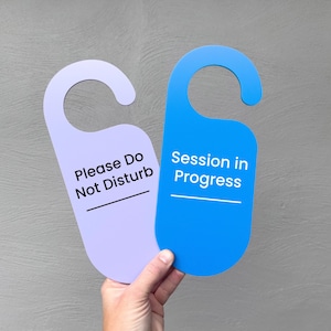 May include: Two door hangers, one lavender and one blue, are held up against a grey background. The lavender hanger reads "Please Do Not Disturb" and the blue one says "Session in Progress."