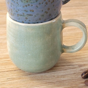 May include: A stack of three handmade ceramic mugs in shades of green and blue. The top mug is a light green, the middle one is blue with brown speckles, and the bottom mug is a muted green. Coffee beans are scattered on the wooden surface.