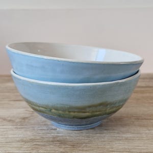 May include: Two handmade ceramic bowls stacked on top of each other. The bowls are light blue on the outside with a gradient of green and gray near the bottom. The inside of the bowls are white.