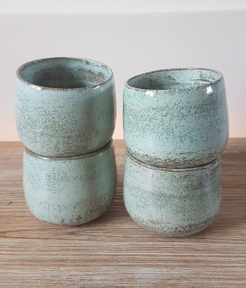 May include: Four ceramic mugs in a light green color with a speckled glaze. The mugs are stacked in pairs.