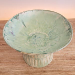 May include: A green ceramic pedestal bowl with a white glaze and a textured base. The bowl has a wide, shallow shape and a smooth, rounded rim.