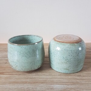 May include: Two ceramic mugs with a speckled light green glaze. The mugs have a rounded shape and a slightly flared lip.