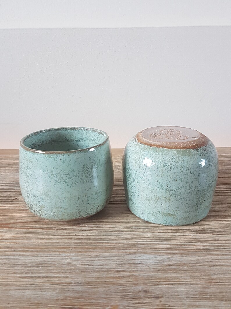 May include: Two ceramic mugs with a speckled light green glaze. The mugs have a rounded shape and a slightly flared lip.