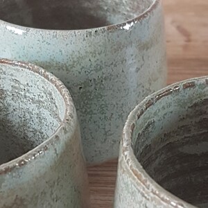May include: Three ceramic mugs with a light green glaze and a speckled finish. The mugs are arranged in a row, with the first two mugs in focus and the third mug out of focus.