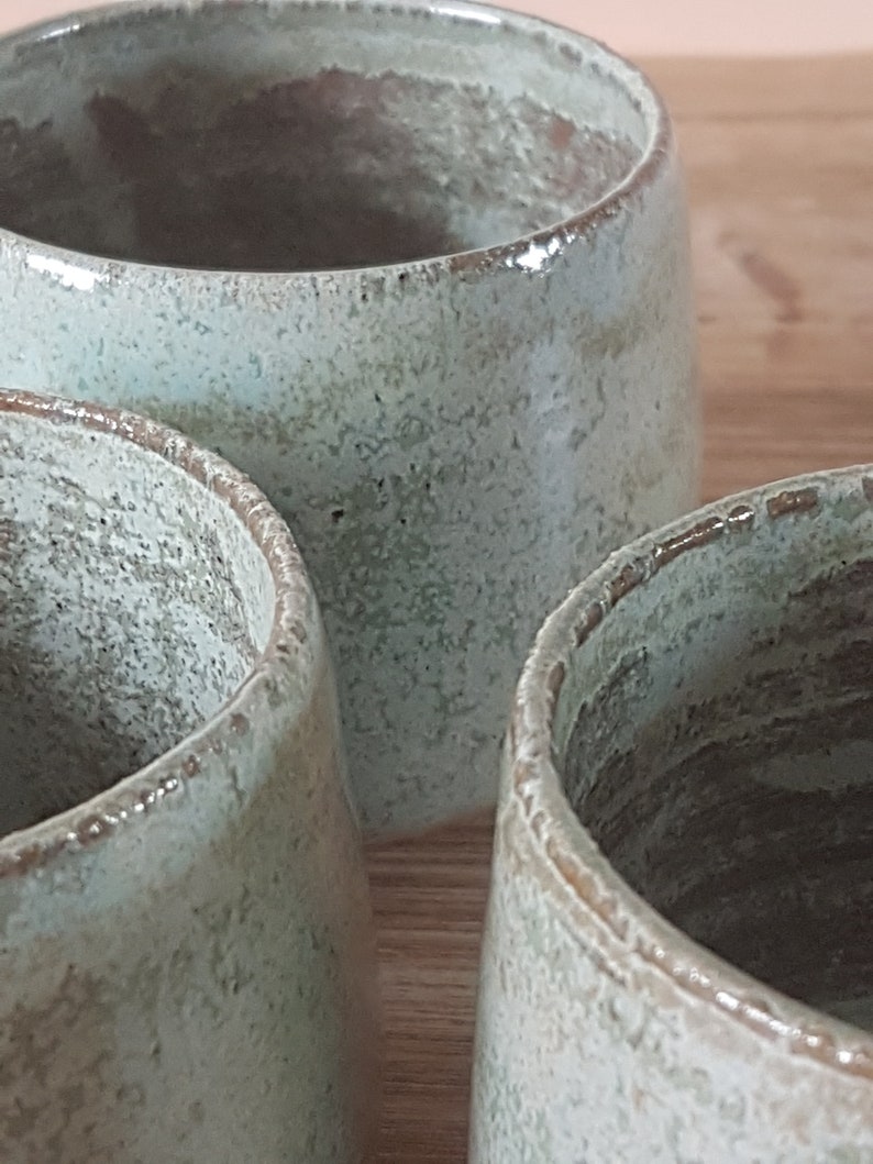 May include: Three ceramic mugs with a light green glaze and a speckled finish. The mugs are arranged in a row, with the first two mugs in focus and the third mug out of focus.