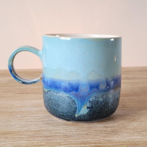May include: A light blue ceramic mug with a dark blue and green glaze. The glaze has a mottled effect, creating a unique and artistic look.