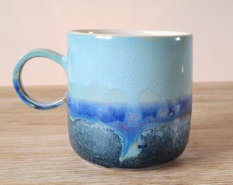 Puede incluir: Una taza de cerámica azul claro con un esmalte azul oscuro y verde. El esmalte tiene un efecto moteado, creando un aspecto único y artístico.