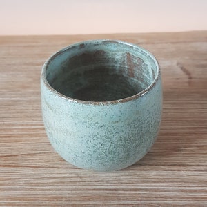 May include: A light blue ceramic cup with a speckled glaze. The cup has a rounded shape and a slightly flared rim.