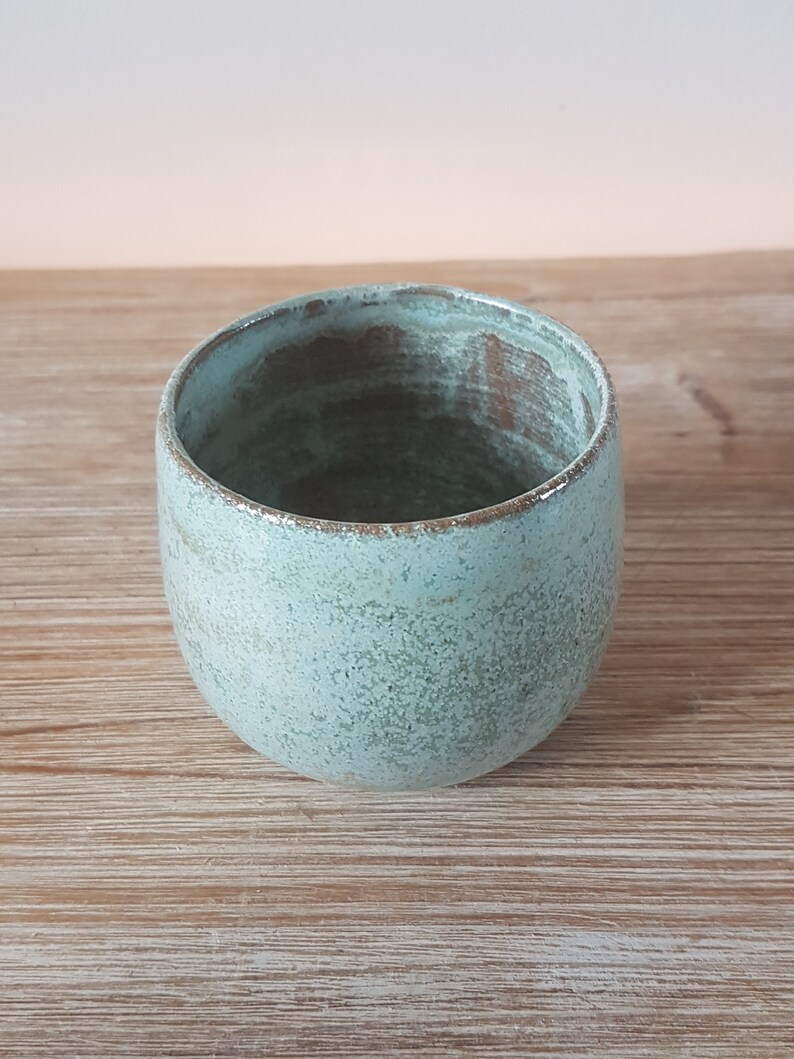May include: A light blue ceramic cup with a speckled glaze. The cup has a rounded shape and a slightly flared rim.