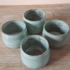 May include: Four teal-colored ceramic mugs with a speckled glaze. The mugs are arranged in a circle on a wooden surface.