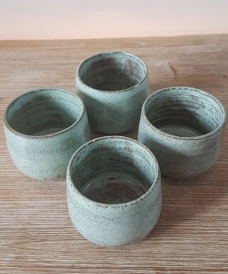 May include: Four teal-colored ceramic mugs with a speckled glaze. The mugs are arranged in a circle on a wooden surface.