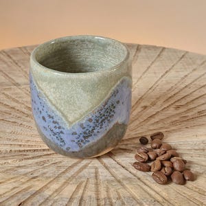 May include: A handmade ceramic coffee cup with a unique color scheme. The cup features a light green top, a blue-lavender center, and a darker green base. A small pile of coffee beans sits next to the cup on a wooden surface.