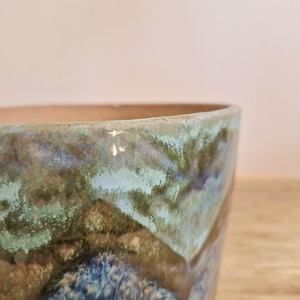 May include: A ceramic planter with a blue, green, and brown glaze. The planter has a wide, round opening and a slightly tapered body.