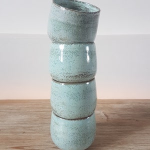 May include: Stack of four light blue ceramic mugs with a speckled glaze. The mugs are stacked on a wooden surface.