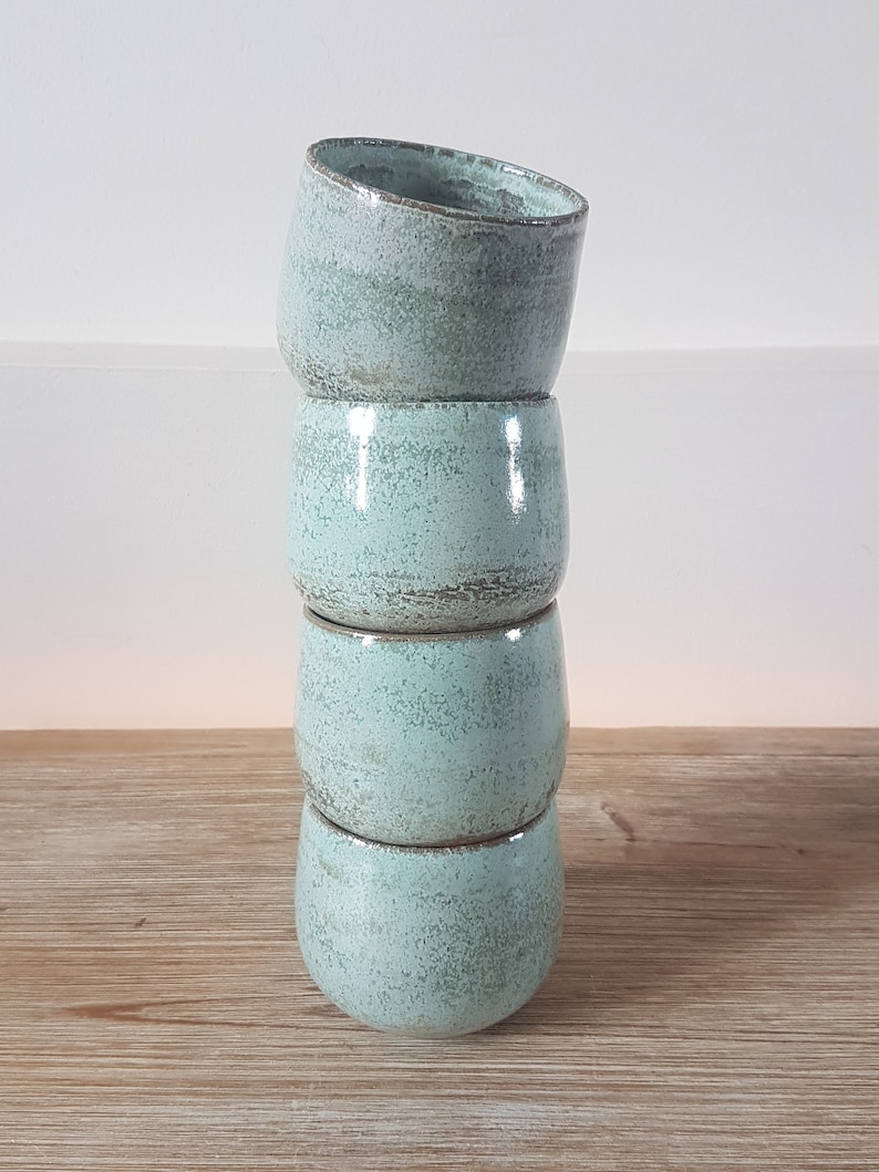 May include: Stack of four light blue ceramic mugs with a speckled glaze. The mugs are stacked on a wooden surface.