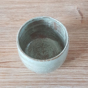 May include: A light blue ceramic cup with a speckled glaze. The cup is empty and sitting on a wooden surface.