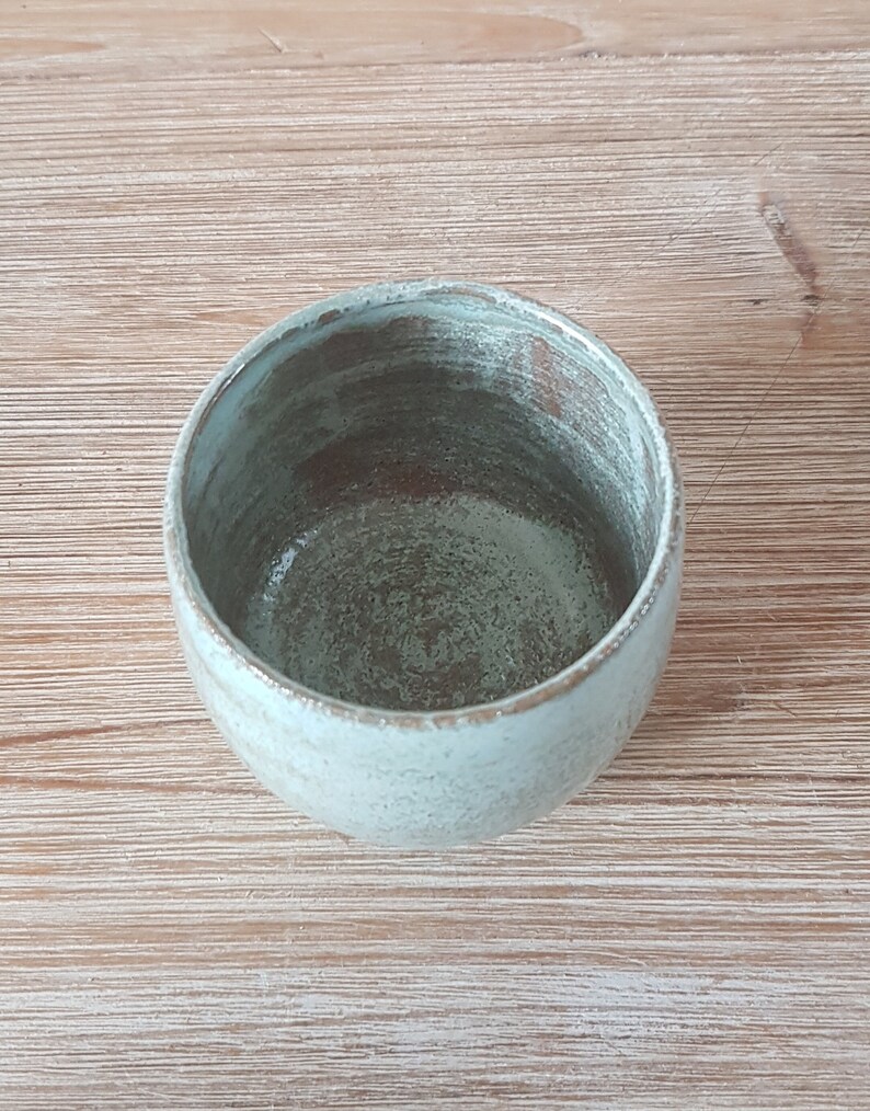 May include: A light blue ceramic cup with a speckled glaze. The cup is empty and sitting on a wooden surface.
