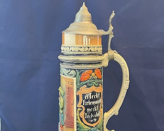 May include: A ceramic beer stein with a pewter lid and handle. The stein is decorated with a colorful scene of a building and flowers. The text "Weckt Erhensmuth werkf Liebshilf" is on the stein.