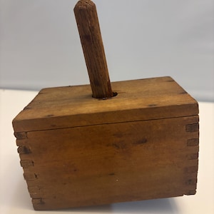May include: A wooden butter mold with a hinged lid and a wooden handle. The mold is made of unfinished wood and has a rustic appearance.