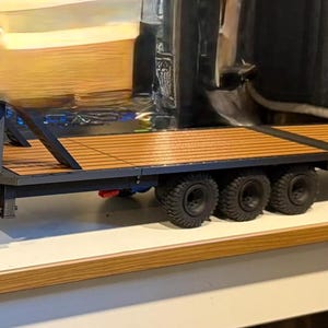 May include: A black toy truck with a trailer. The trailer has a wooden deck and three sets of tyres. The truck is connected to the trailer. The truck is black.