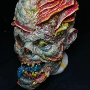 May include: A grotesque, detailed mask of a zombie-like creature. The mask features a decaying, green and yellow complexion with exposed teeth, a blue tongue, and red and orange fungal growths on the head. The eye is a vibrant green.
