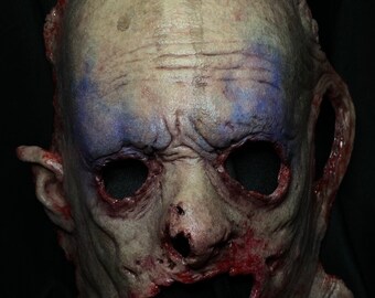 May include: A realistic horror mask with a pale, fleshy face, bloody wounds, and blue paint around the eyes. The mask has a wide, open mouth with a bloody, fleshy interior.