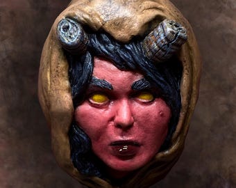May include: A red-skinned, female figure with black hair and yellow eyes is depicted in a clay sculpture. The figure is wearing a brown hooded cloak and has horns on her head.